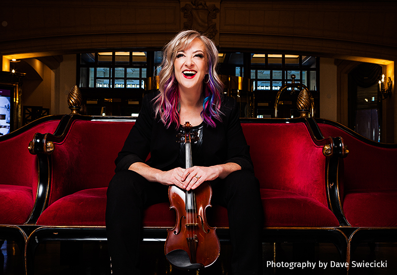 Karen Barg smiling on red velvet couch with violin.